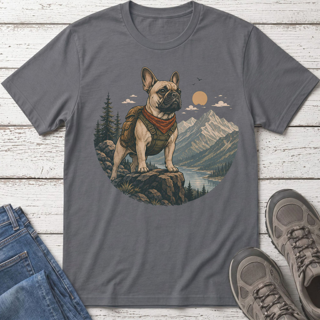 French bulldog wearing backpack and bandana stands on rocks overlooking mountains and lake.