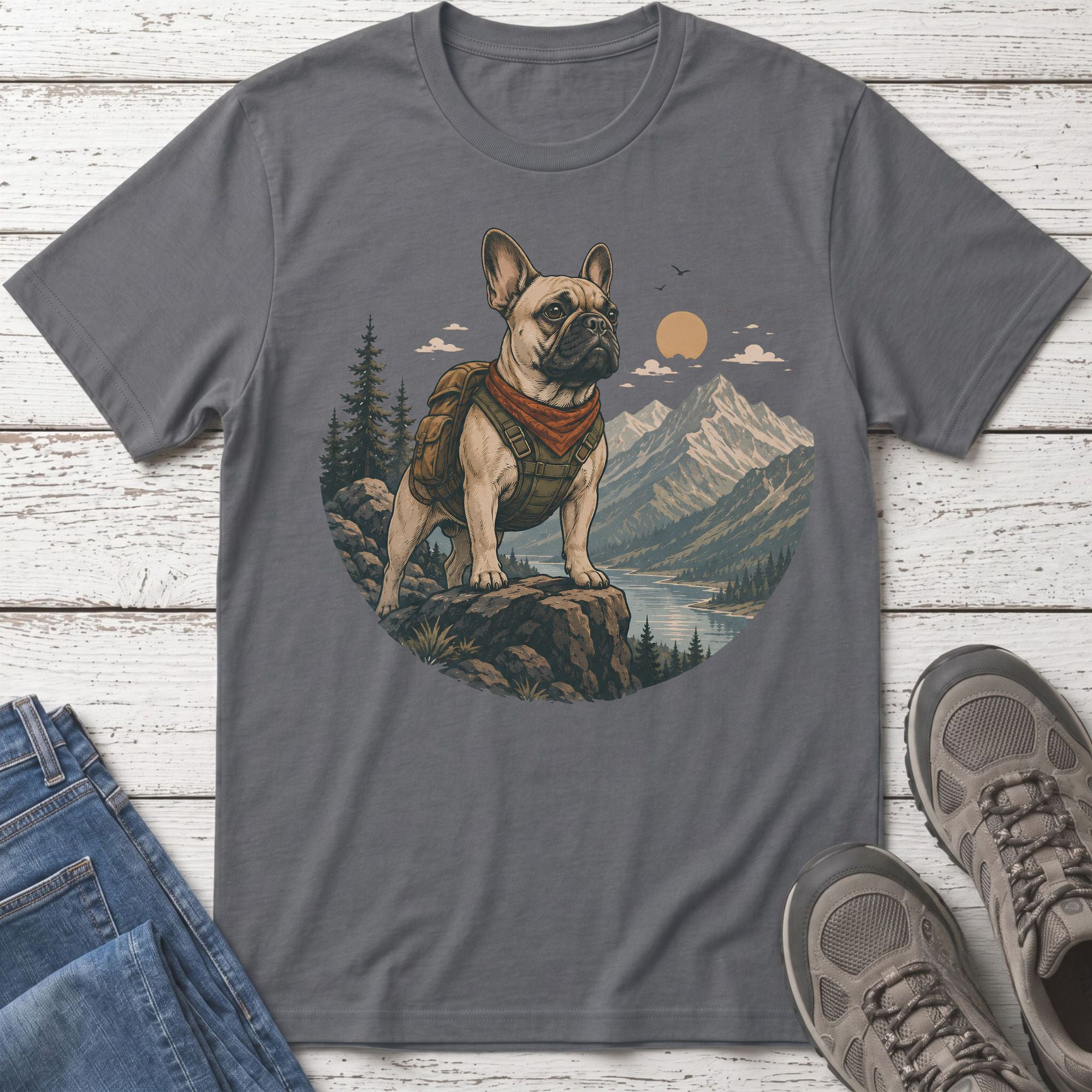 French bulldog wearing backpack and bandana stands on rocks overlooking mountains and lake.