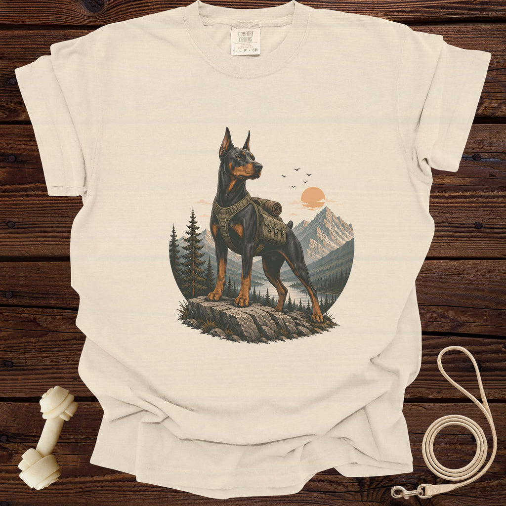 Doberman wearing a backpack on a rocky outcrop with mountains and lake.