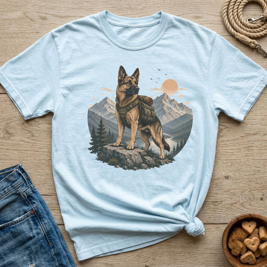 German Shepherd wearing a backpack stands on a rock with mountains and a river behind it.