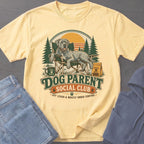 Yellow t-shirt with a graphic of a dog on a chair, trees, and the text "Dog Parent Social Club".