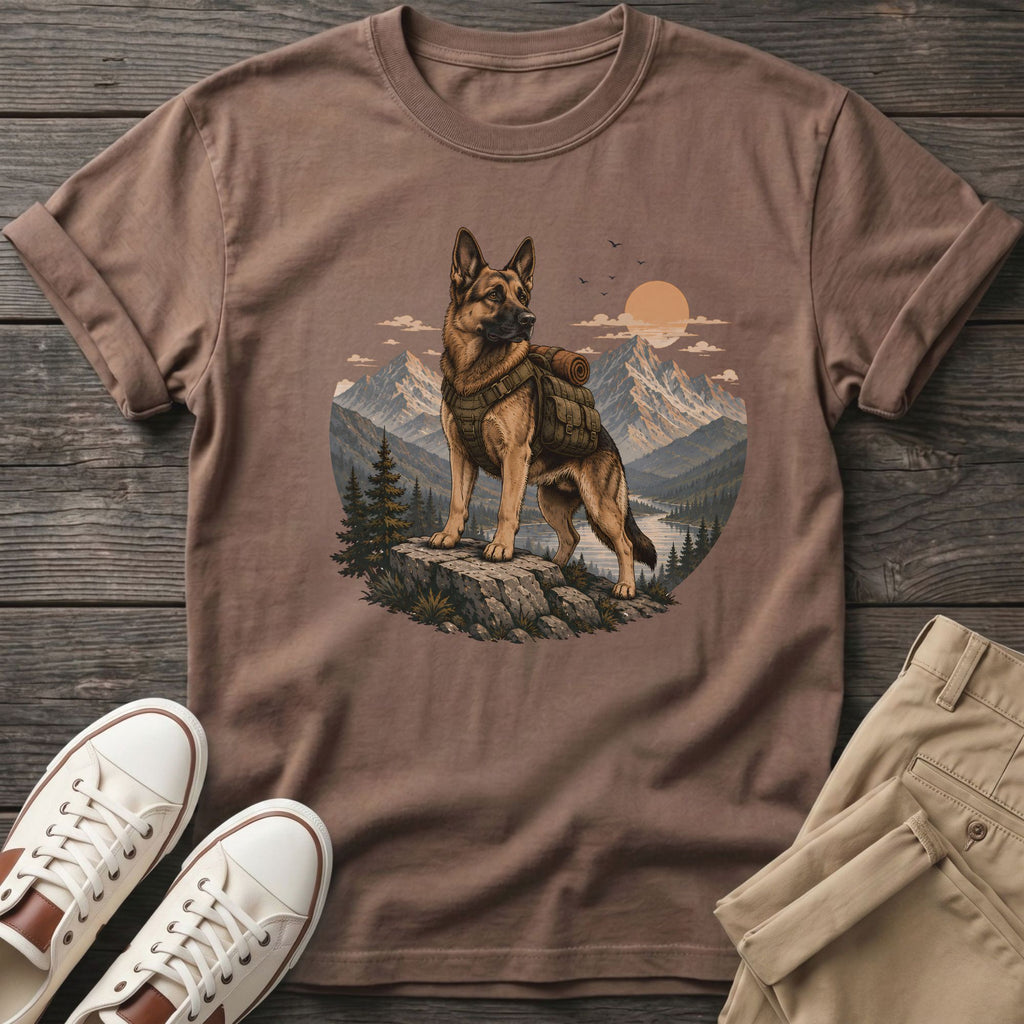 German Shepherd wearing a backpack stands on a rock with a mountain landscape background.