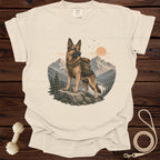 German Shepherd wearing a backpack stands on a rock in a mountainous landscape.