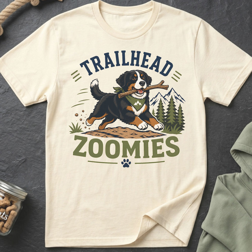 Bernese Mountain Dog with stick, text "TRAILHEAD ZOOMIES," mountains, and trees graphic on a cream t-shirt.