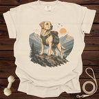 Labrador retriever wearing a backpack stands on a rock overlooking a lake and mountains.