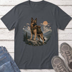 German Shepherd wearing a backpack stands on a rock with mountains and a river behind.