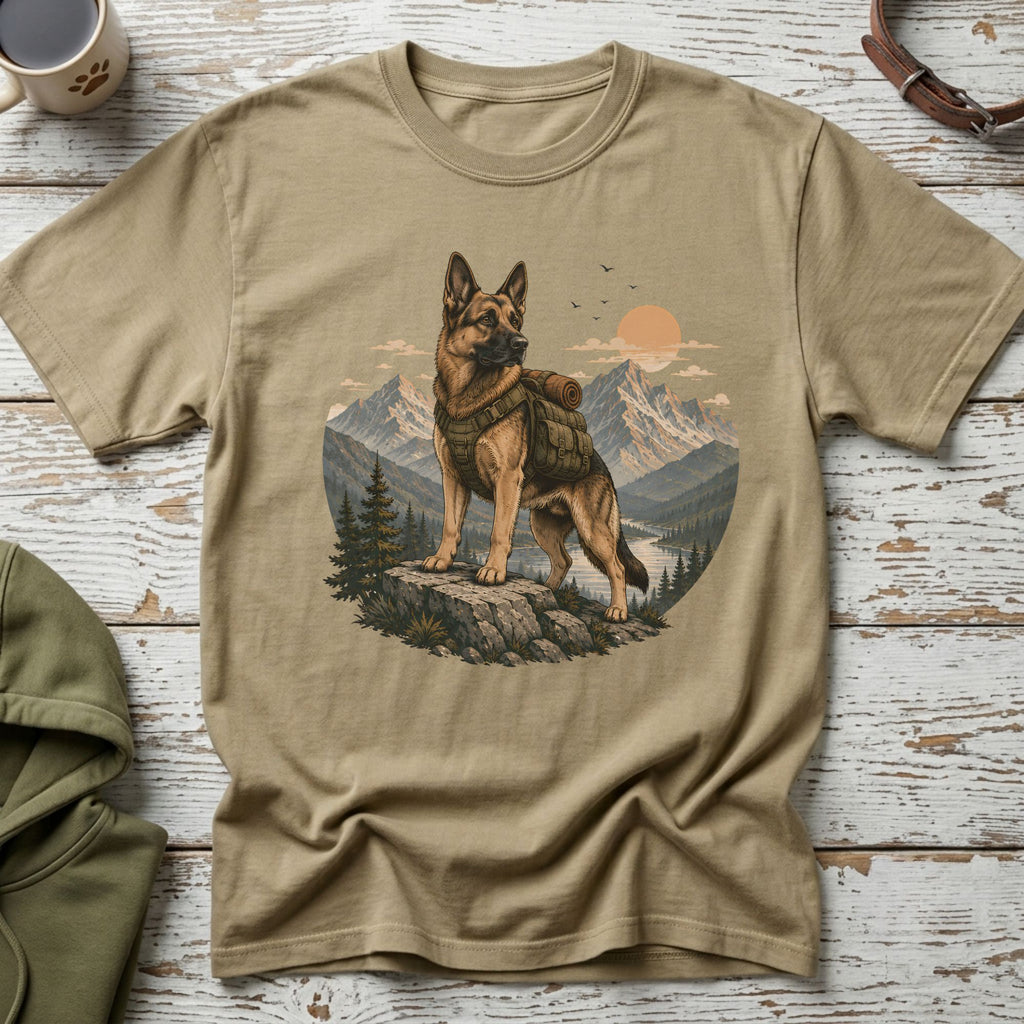 German Shepherd wearing a backpack stands on rocks with mountains and river behind.