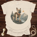 French bulldog wearing a backpack and bandana stands on a cliff overlooking mountains and a lake.
