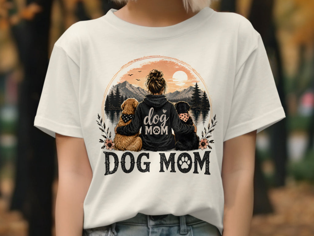 White t-shirt with "Dog Mom" design featuring a woman and two dogs by a lake at sunset.