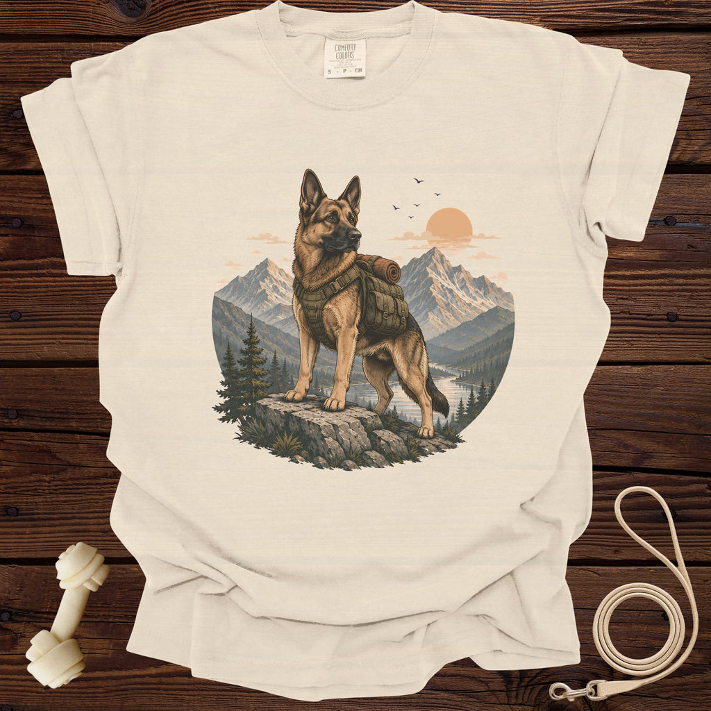 German Shepherd with backpack stands on rocks overlooking mountains and river at sunset.