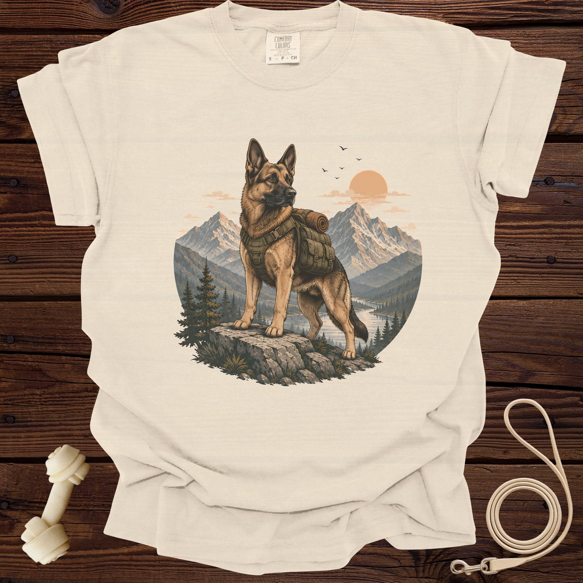 German Shepherd with backpack stands on rocks overlooking mountains and river at sunset.