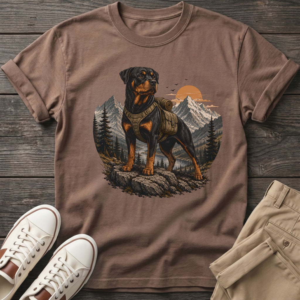 Rottweiler wearing a backpack stands on rocks with mountains, trees, and sunset behind.