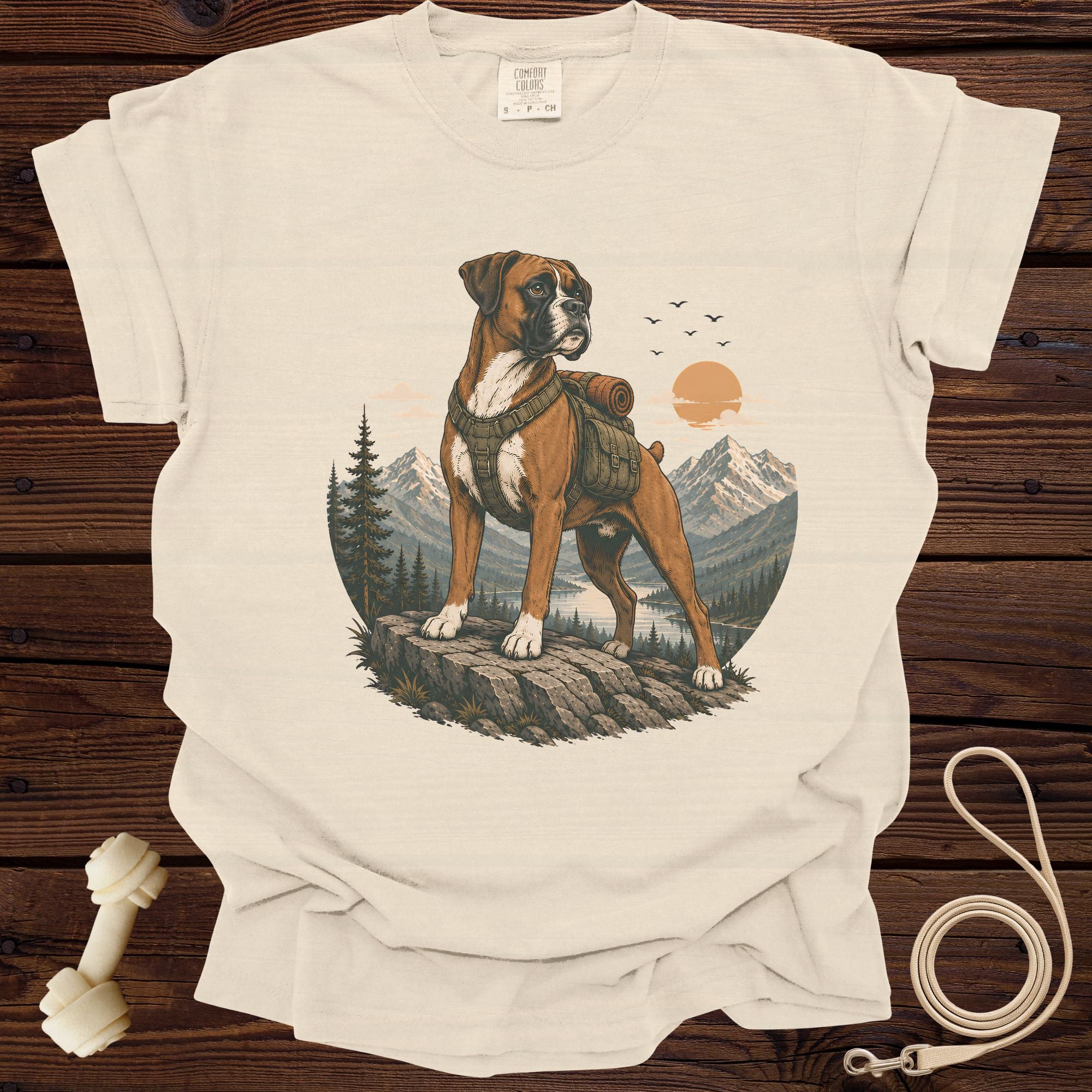 T-shirt graphic featuring a boxer dog wearing a backpack on a rocky cliff with mountains and a lake.